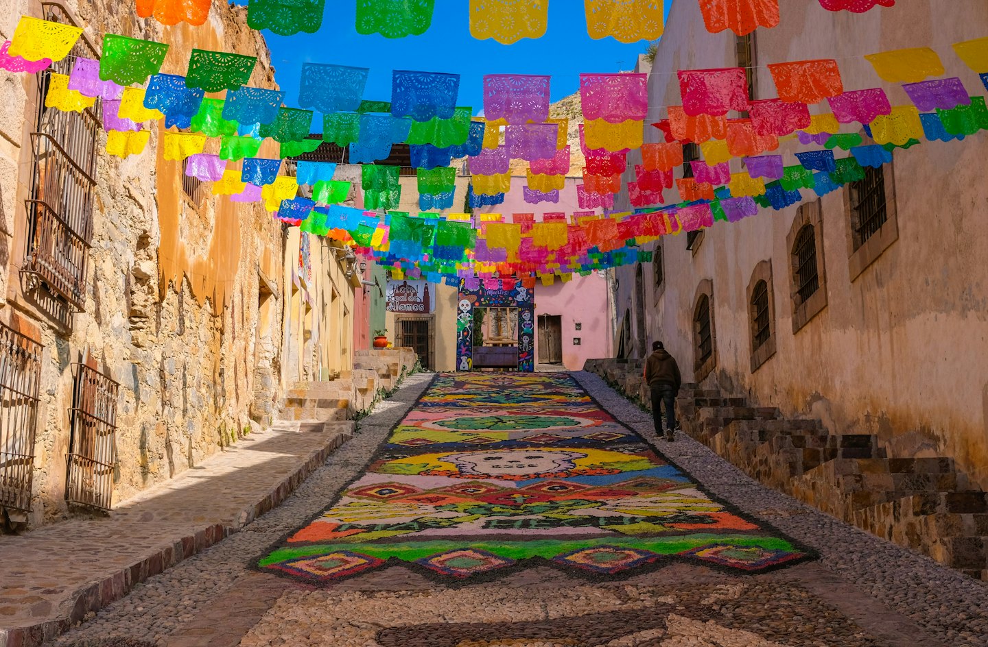 Persona camina por un callejón de adoquines decorado con arte de arena de colores.
